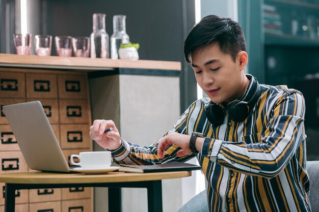 Young man sitting with a laptop in cafe and looking at his watch attentively