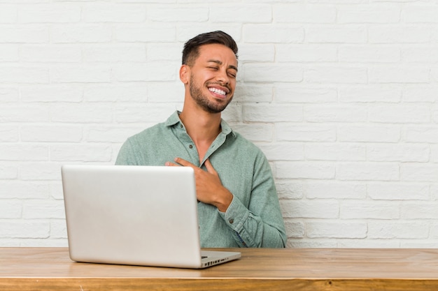 Young man sitting working with his laptop laughs out loudly keeping hand on chest