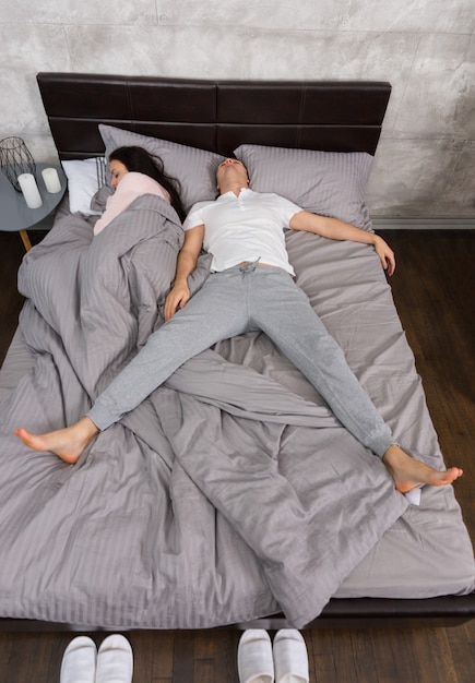 Young man sleeping in free fall position with his wife occupied the whole bed, wearing pajamas, near bedside table with candles