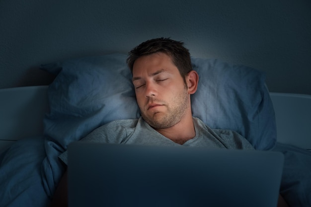 Foto giovane che dorme davanti a un portatile in un letto a casa stanco dopo la giornata di lavoro maschio si addormenta mentre