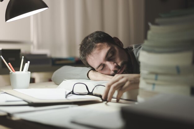 Photo young man sleeping at his desk late at night he is leaning on a book and holding glasses stress and exhaustion concept