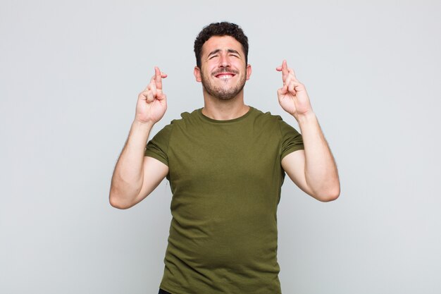 Young man smiling and anxiously crossing both fingers, feeling worried and wishing or hoping for good luck