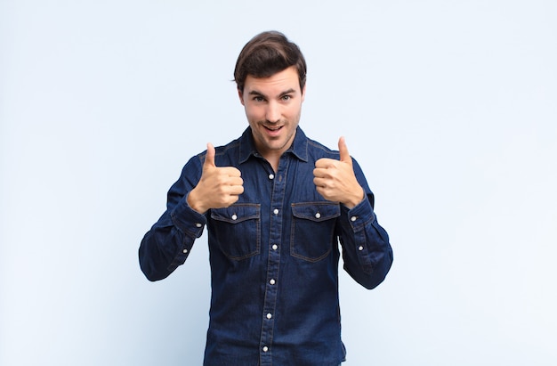 Young man smiling broadly looking happy, positive, confident and successful, with both thumbs up on blue wall