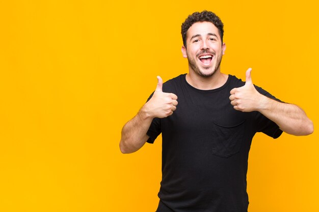 Young man smiling broadly looking happy, positive, confident and successful, with both thumbs up