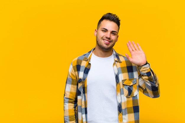 Young  man smiling happily and cheerfully, waving hand, welcoming and greeting you, or saying goodbye against orange wall