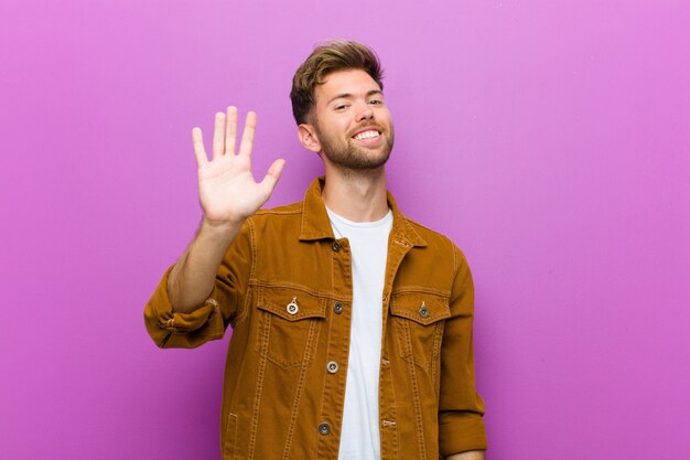 Young man smiling happily and cheerfully, waving hand, welcoming and greeting you, or saying goodbye against purple wall