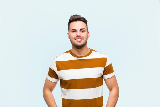 Young man smiling happily with a hand on hip and confident, positive, proud and friendly attitude over blue wall