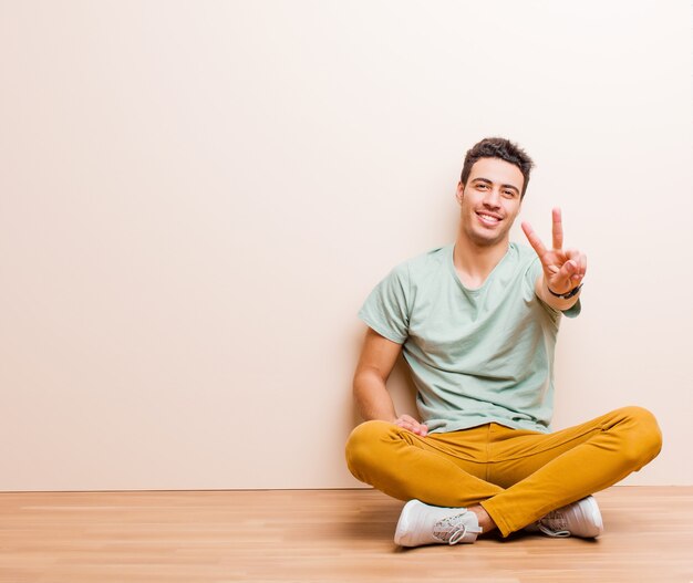 Young man smiling and looking friendly, showing number two or second with hand forward, counting down sitting on the floor