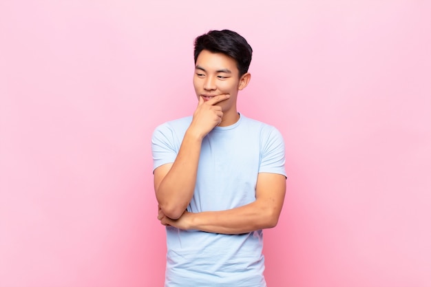 Young man smiling with a happy, confident expression with hand on chin, wondering and looking to the side over wall