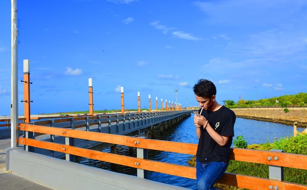 Young man smoking cigarette on bridge against blue sky