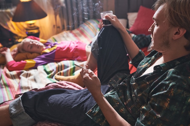 Young man smoking a cigarette and drinking wine sitting on the bed with his girlfriend relaxing near by him at home