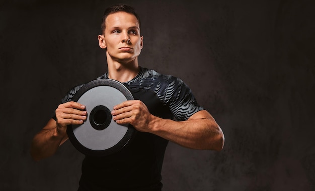 A young man in sportswear holding a barbell disk on a dark background.