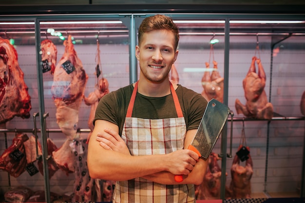 Photo young man stand at meat shelf in grocery store. he hold butcher knife in hand and pose. animal brawn behind. positive male worker smile.