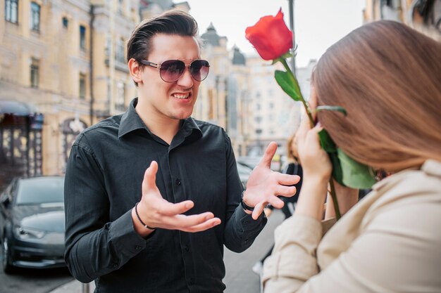 Young man stand outside in front of woman and talk to her. She holds red rose in hands. Guy wear sunglasses.