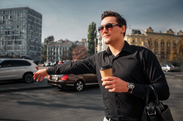 Young man stand outside and hold cup of drink. He holds hand. Guy try to catch taxi.