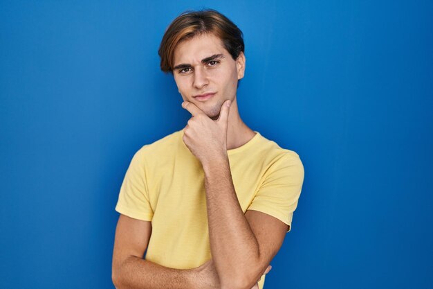 Young man standing over blue background looking confident at the camera with smile with crossed arms and hand raised on chin thinking positive