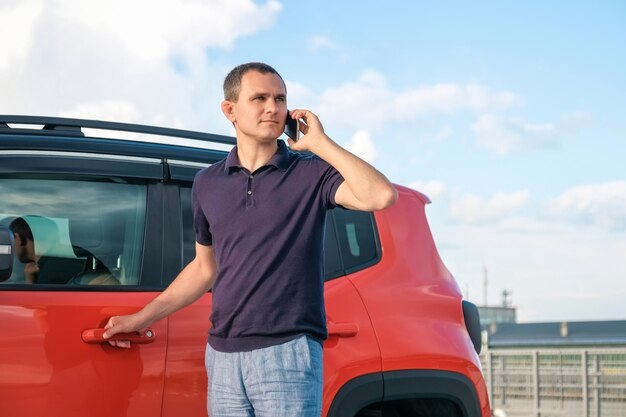 A young man stands near the car and speaks on the phone outdoors