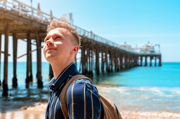 A young man stands near a pier on Malibu beach in California