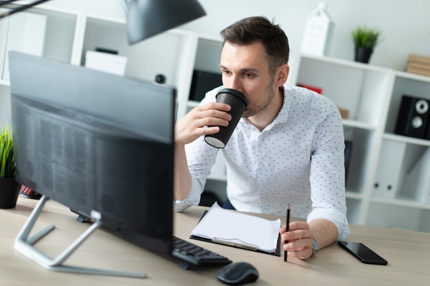 A young man stands near a table in the office, holds a pencil in his hand and drinks coffee. A young man works with documents and a computer.