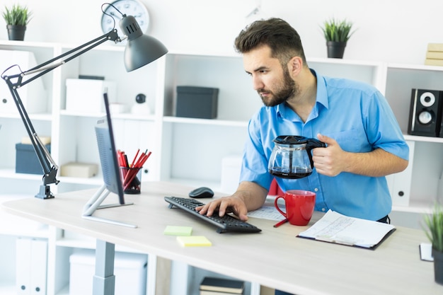 若い男がコンピューターテーブルのオフィスに立ち、コーヒーをbrewれています。