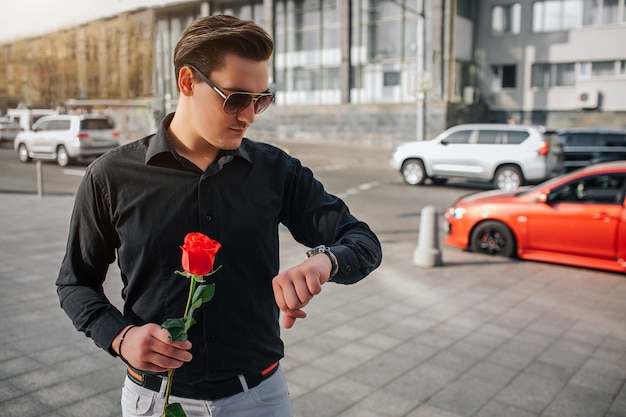 Young man stands outside on street and hold red rose. He looks at watches. Guy is waiting. There is road with cars behind him.