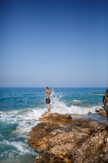 Un giovane si erge sulle rocce che si affacciano sul mar mediterraneo aperto. un ragazzo in una calda giornata di sole estivo guarda la brezza marina
