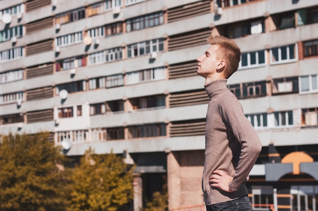Молодой человек с закрытыми глазами стоит перед многоэтажным домом.