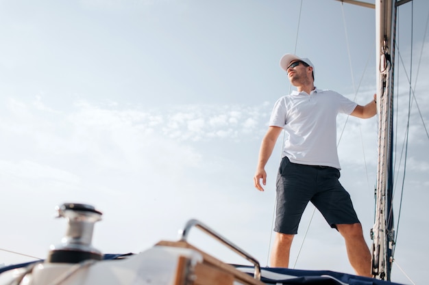 Il giovane sta sul bordo dell'yacht e guarda in avanti. tiene sull'albero con la mano. il giovane pone. indossa camicia bianca e pantaloncini neri.