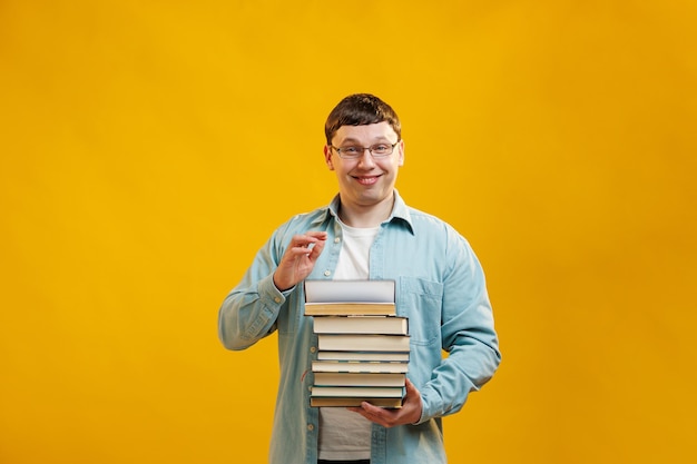 Young man student in glasses holds stack of university books from college library on yellow background Happy guy smiles he is happy to graduate education abroad concept Copy space