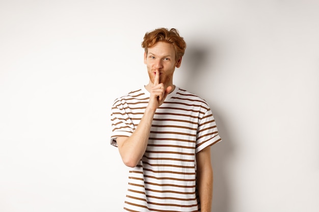 Young man student with red hair shushing, telling a secret and smiling, standing over white background.