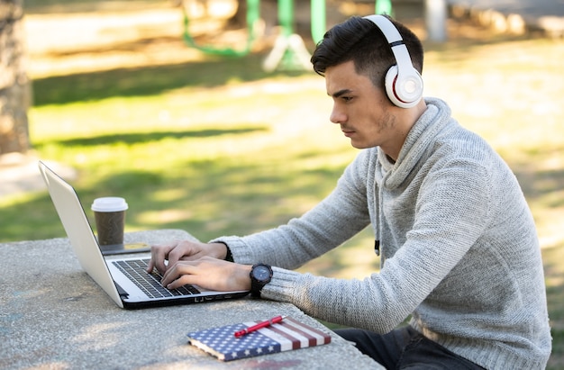 マスクなしで公園でヘッドフォンで音楽を聴きながらコンピューターで勉強している若い男