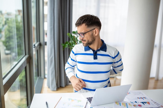 Young man suffering from shoulder and back pain while sitting in office and working on laptop Business man stretching while working on laptop Stressed businessman suffering from neck pain