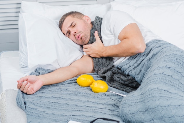 레몬과 온도계와 함께 침대에 누워 목의 통증으로 고통받는 젊은 남자