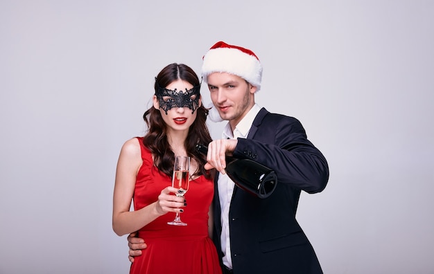 Young man in suit and christmas hat and woman in a red dress and mask holding champagne glass