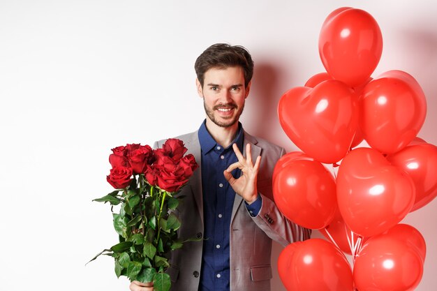 スーツを着た若い男は、OKのサインを示し、赤いバラを持って、バレンタインデー、白い背景にハートの風船と花でロマンチックな驚きを準備し、すべてを制御しています。