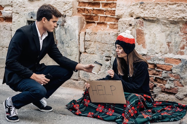 A young man in a suit sat down on the street floor with beggars and gives a cup of coffee