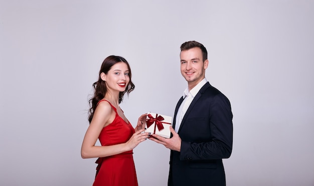 Young man in suit and woman in a red dress holding a gift box together isolated on gray background