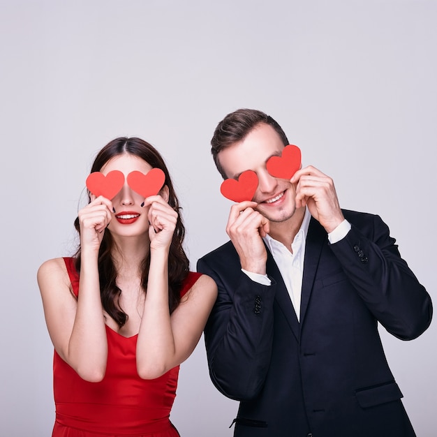 Young man in suit and woman in red dress holding red hearts over their eyes on white background