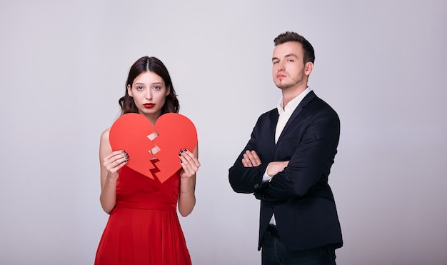 Young man in suit and woman in red dress holds a red broken heart on white background