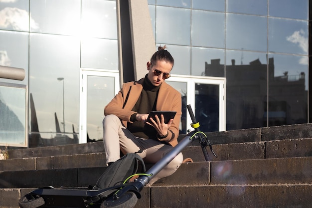 Young man on sunglasses sitting remote working on the tablet sending and responding emails with an electric scooter