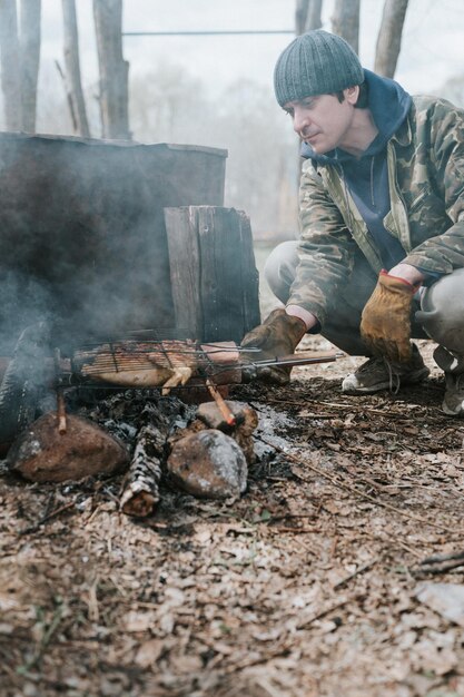 Young man survivalist cooks roasts chicken meat food are fried on grill on smoldering coals or ember from a campfire on the ground barbecue in camping conditions countryside and wild rustic life