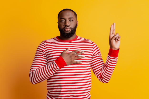 Young man swearing with hand on chest and fingers up serious face on yellow wall