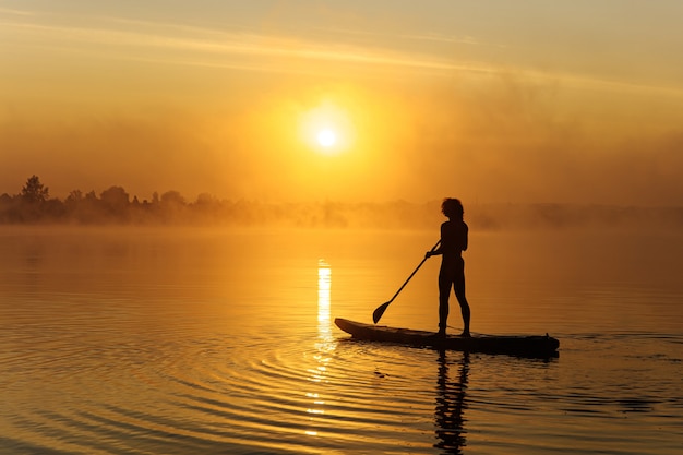 Young man in swimming trunks doing surfing on sup board during amazing sunrise at local lake.