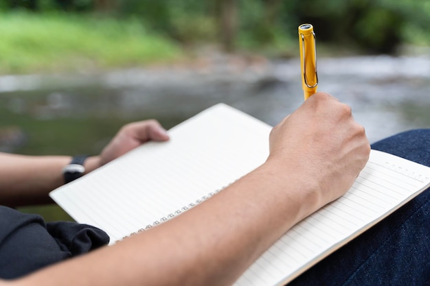 Young man takes notes in a notebook on nature Man sits by a tree with his back to him a pencil and a notebook in his hands takes notes