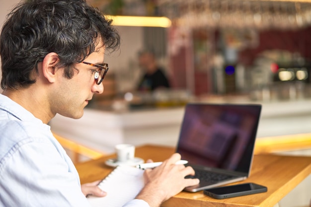 Young man takes notes while working on laptop at cozy cafe