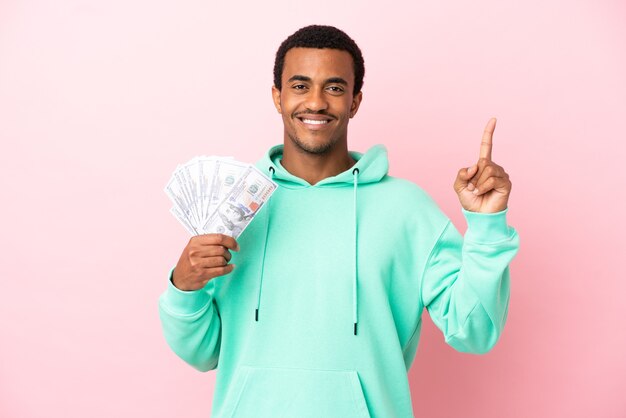 Young man taking a lot of money over isolated pink background pointing up a great idea