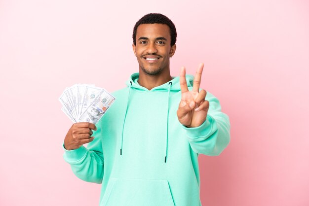 Young man taking a lot of money over isolated pink background smiling and showing victory sign