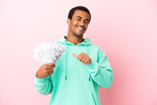 Young man taking a lot of money over isolated pink surface proud and self-satisfied