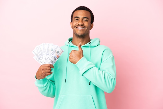 Young man taking a lot of money over isolated pink wall giving a thumbs up gesture