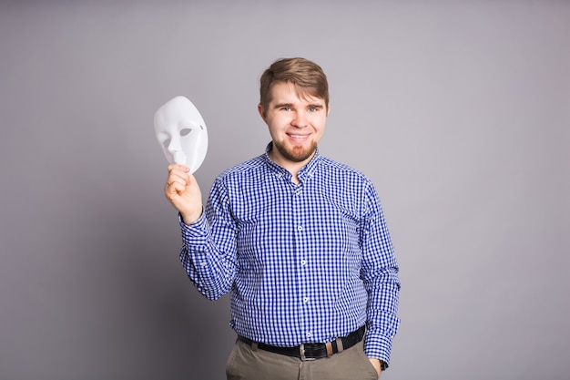 Young man taking off plain white mask revealing face, gray\
wall